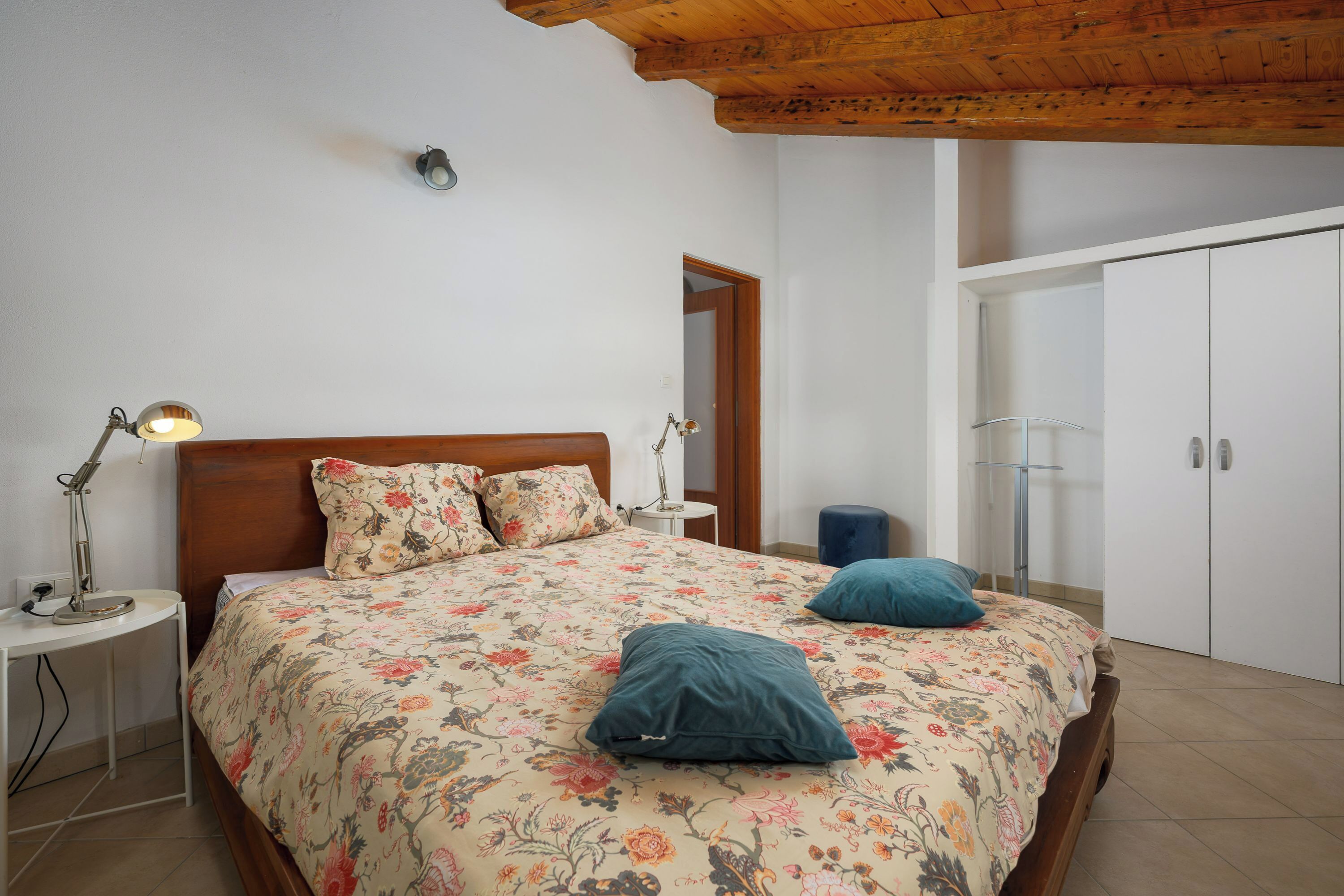 A bedroom with a wooden ceiling, a bed with floral bedding, bedside lamps, a wardrobe, and a small table.
