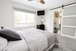 Ein Schlafzimmer mit Bett, Fenster, Fernseher, Schreibtisch und einer Schiebetür zum Badezimmer.