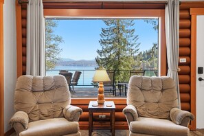 A room with two cushioned chairs, a small table with a lamp, and a large window offering a view of a lake and forest.