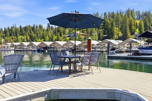 A wooden deck with a table and chairs, a large umbrella, and a row of covered structures along a body of water.
