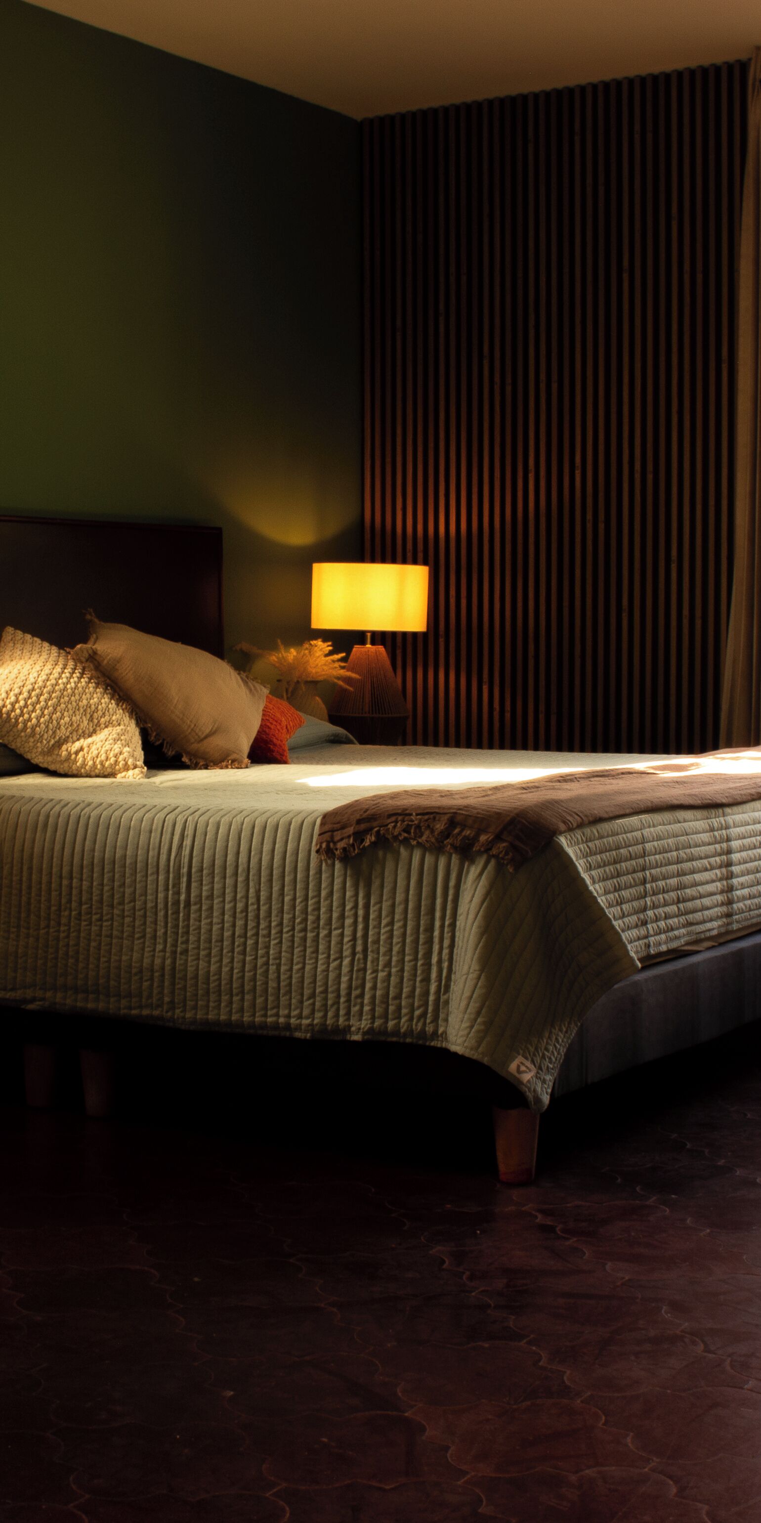 A neatly made bed with a lamp beside it, a dark curtain in the background, and a patterned carpet.