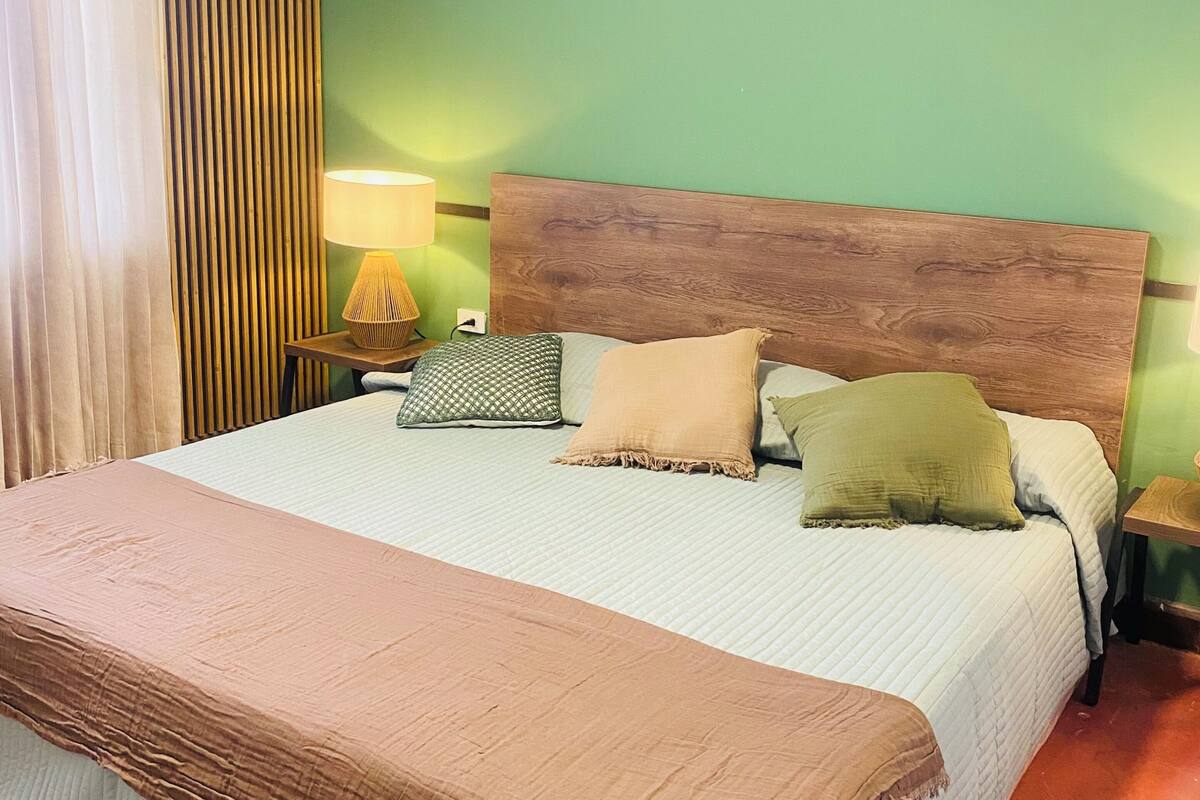 A hotel room with a large bed, a wooden headboard, a nightstand with a lamp, and a green wall.