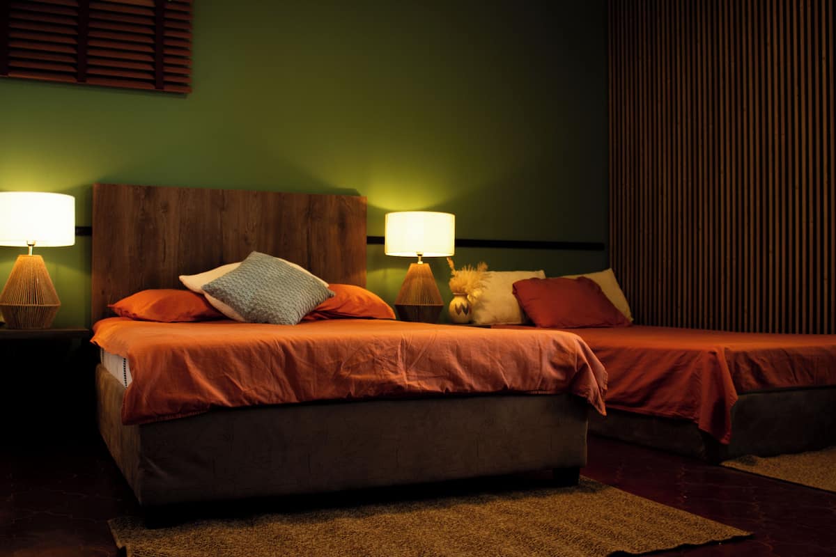 A bedroom with two beds, each with a wooden headboard, two bedside lamps, and a green wall.