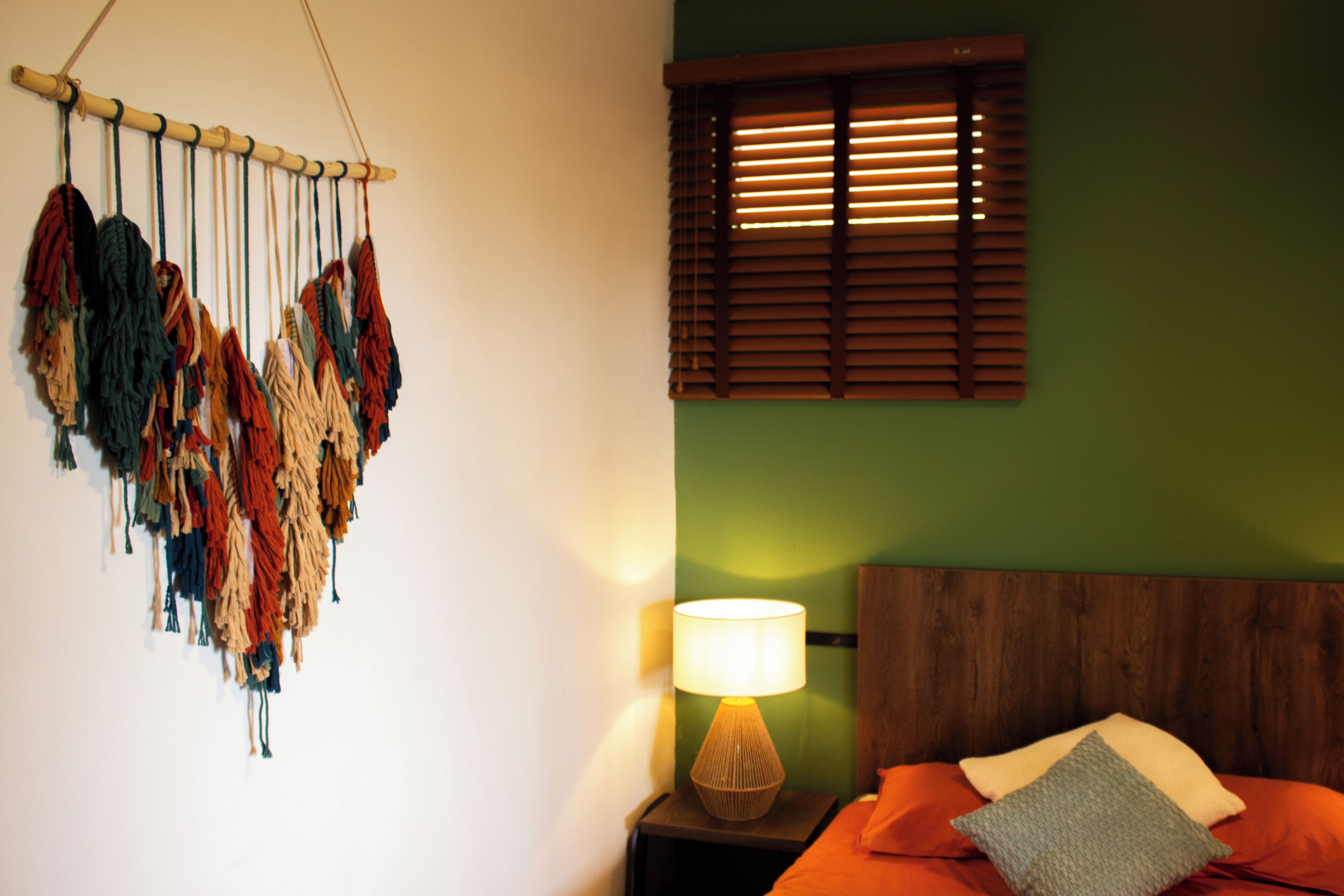 Una habitación con una cama, una lámpara de pared, una ventana con persianas y un cuadro colgado.