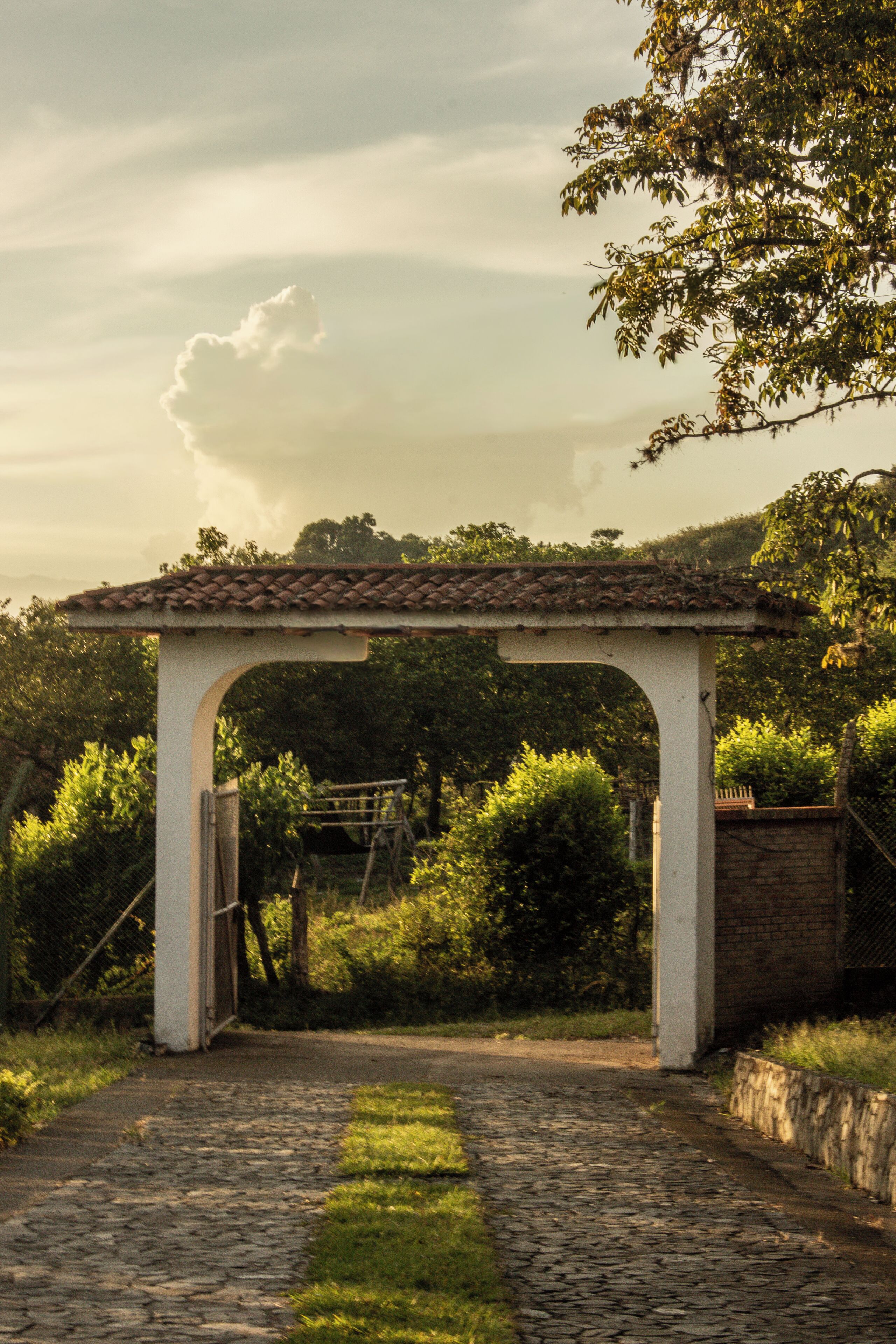 A stone pathway leading to a white pergola with a tiled roof, surrounded by greenery.