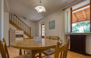 A dining area with a round wooden table, chairs, a staircase, a radiator, and a window with a view of greenery.