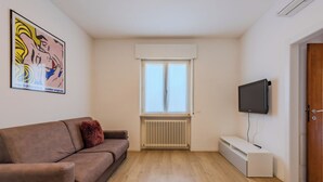 A living room with a brown sofa, a television mounted on the wall, a radiator, and a window with white curtains. - Riva Olive by Interhome (Riva del Garda)