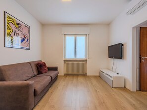 A living room with a brown sofa, a television mounted on the wall, a radiator, and a window with white curtains. - Riva Olive by Interhome (Riva del Garda)