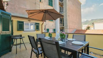 Zona pranzo con tavolo apparecchiato per due, sedie da esterno, ombrellone e piante in vaso.