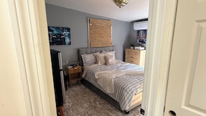 A bedroom with a bed, a nightstand, a dresser, a wall-mounted air conditioner, and a ceiling light fixture.