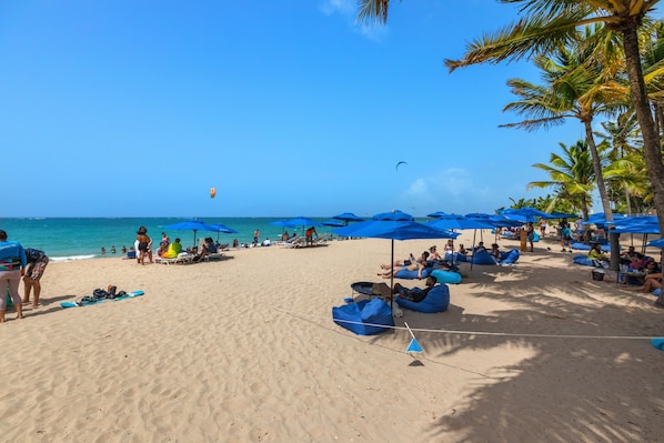 A beach with blue umbrellas, lounge chairs, and people relaxing. - Coastal Summer Villa: Pool/Kitchen/Patio (San Juan)