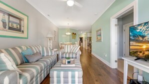 A living room with a striped sofa, ottoman, and a television on a stand. - Residence 103N at The Sandcastle Condominiums (Wildwood Crest)