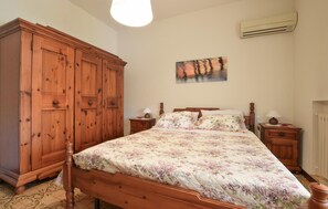 A bedroom with a wooden wardrobe, a bed with floral bedding, two bedside tables, and an air conditioning unit on the wall.