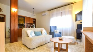 A living room with a sofa, a blue armchair, a round wooden table, and a kitchen area with cabinets and a stove.