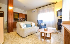 A living room with a sofa, a blue armchair, a round wooden table, and a kitchen area with cabinets and a stove.