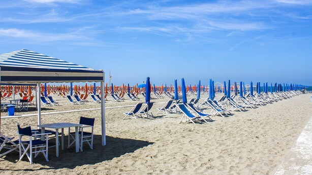 A beach with rows of sun loungers and umbrellas, a covered seating area, and a clear blue sky.