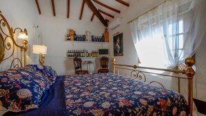 A bedroom with a bed, two chairs, a desk with a computer, shelves with bottles, and a window with curtains. - La Duchessa Tenuta Donna Olga (Porrona)