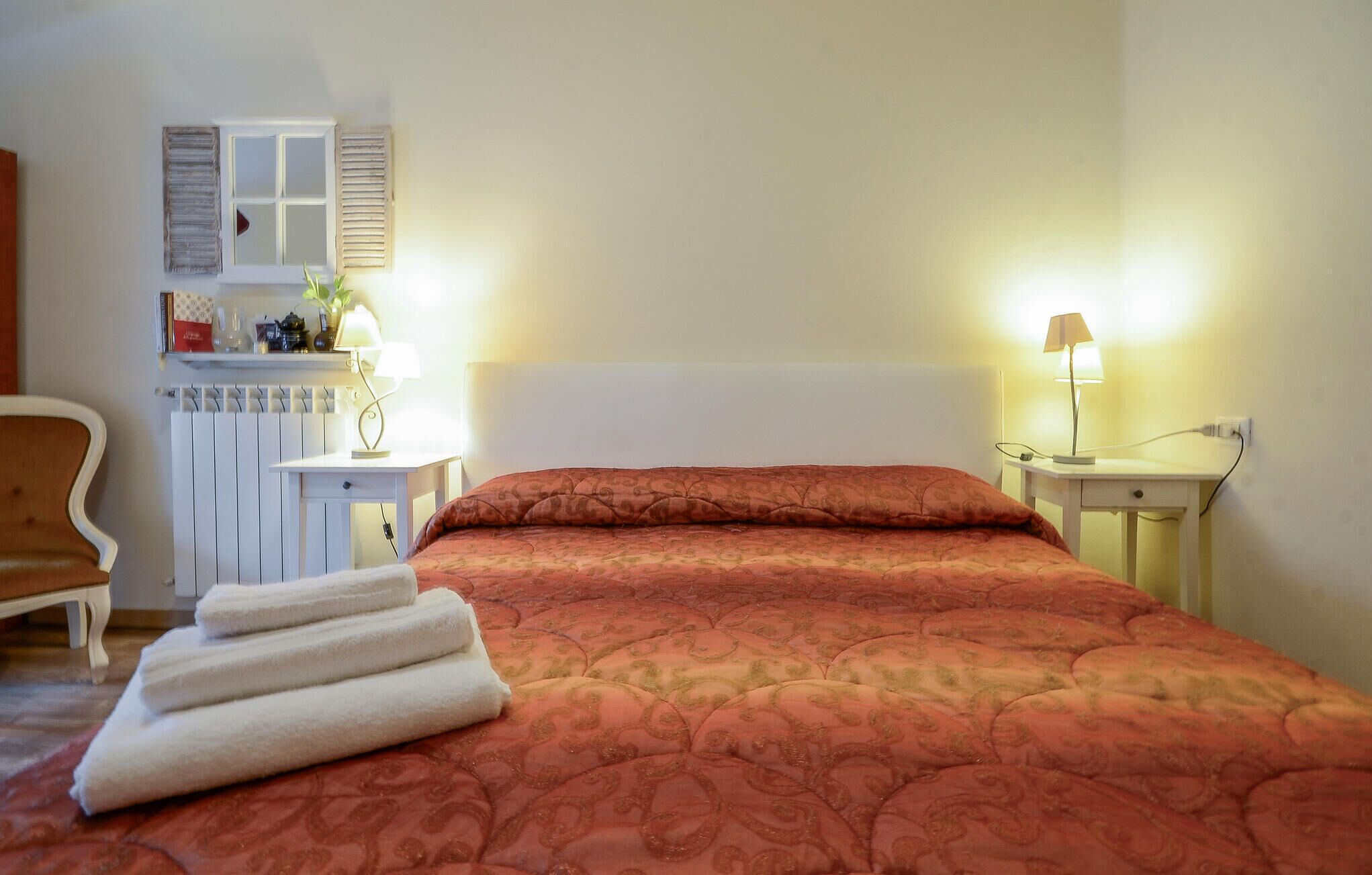 A neatly made bed with a patterned bedspread, a bedside lamp, a chair, and a radiator.