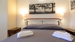 Un lit bien fait, recouvert d’une couverture grise et agrémenté d’oreillers, deux tables de chevet avec des lampes, et un tableau représentant des arbres accroché au mur.