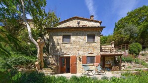 Une maison en pierre avec une porte en bois, un petit balcon et un jardin agrémenté de plantes et d’une chaise.