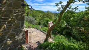 Un mur de pierres surmonté d’un portail en métal, un sentier pavé de pierres et une vue sur un paysage verdoyant, avec des arbres et des collines à l’horizon.