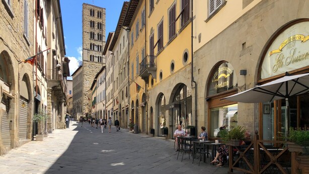 Une rue historique bordée d’immeubles, avec une tour imposante et des tables installées à l’extérieur d’un café.