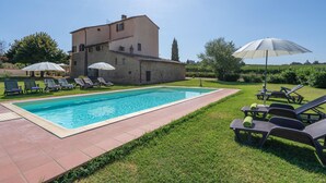 Une piscine avec des chaises longues et des parasols, un bâtiment en pierre et une pelouse.