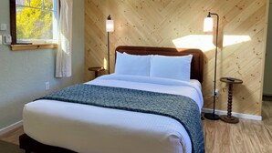 Un dormitorio con pared de madera en patrón de espiga, una cama con ropa de cama blanca, dos lámparas de noche, una ventana con cortinas y una mesita redonda pequeña.
