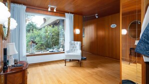 Quarto com painéis de madeira, uma janela grande com cortinas, uma poltrona branca e piso de madeira.