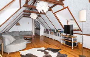 Ein Loft-Zimmer mit Holzbalkendecke, einer Couch, einem Fernseher, einem Bücherregal und einem Kuhfellteppich.