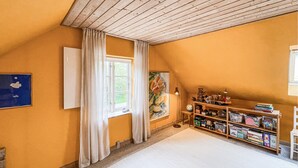 Ett rum med ett sluttande tak, ett fönster med gardiner, en bokhylla och en tavla på väggen.