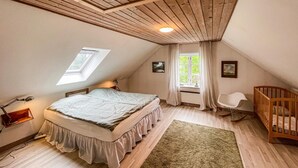 Ett sovrum med ett sluttande tak, en säng, en spjälsäng, ett fönster med gardiner och ett litet skrivbord med en lampa.