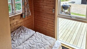 A wooden cabin room with a bed, a window, and a view of a barbecue grill outside.