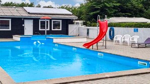 Una piscina rectangular con un tobogán rojo, un pequeño edificio con techo de tejas y una zona de terraza pavimentada con sillas de plástico.
