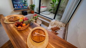 Ein Holzesstisch mit gewebten Tischsets, eine Obstschale und eine Blumenkomposition. Ein Fernseher ist an der Wand befestigt, und durch eine Glastür ist eine Badewanne zu sehen.