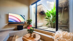 Ein modernes Zimmer mit großem Fernseher, einem Holztisch mit Blumenvase und Blick auf einen Bereich mit Strohdach im Freien.