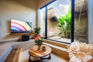 Ein modernes Zimmer mit großem Fernseher, einem Holztisch mit Blumenvase und Blick auf einen Bereich mit Strohdach im Freien.
