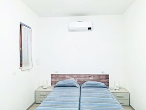 Un dormitorio con una cama, dos mesitas de noche, una lámpara de noche, un ventanal con cortinas y un equipo de aire acondicionado.