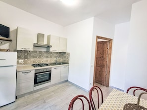 A compact kitchen with a gas stove, oven, and white cabinetry. A dining area with a patterned table and chairs is visible. - Wohnung mit Wasserblick by Interhome (Capoliveri)