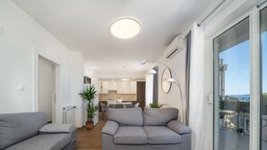 Un apartamento moderno con dos sofás grises, una mesa de centro blanca, una cocina al fondo y un balcón con vistas.