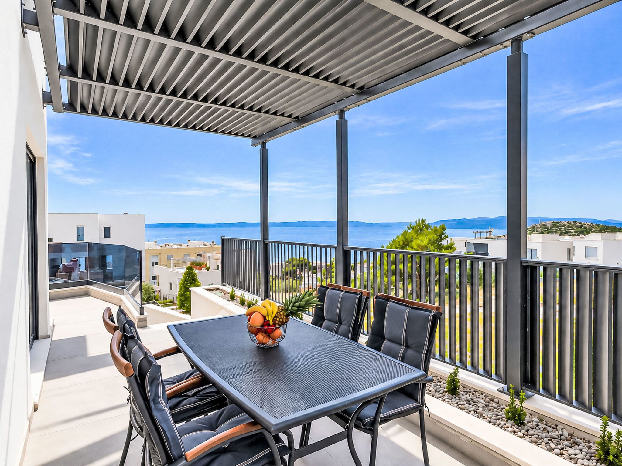 A covered outdoor seating area with a table and chairs, overlooking a body of water and distant hills.