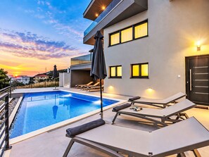 Et moderne hotel med swimmingpool, liggestole og havudsigt i solnedgang.