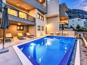 A modern outdoor swimming pool with lounge chairs and umbrellas, surrounded by a building with balconies and outdoor lighting. - Villa Vip in Makarska mit beheiztem Pool und 4 Schafzimmern by Interhome (Makarska)