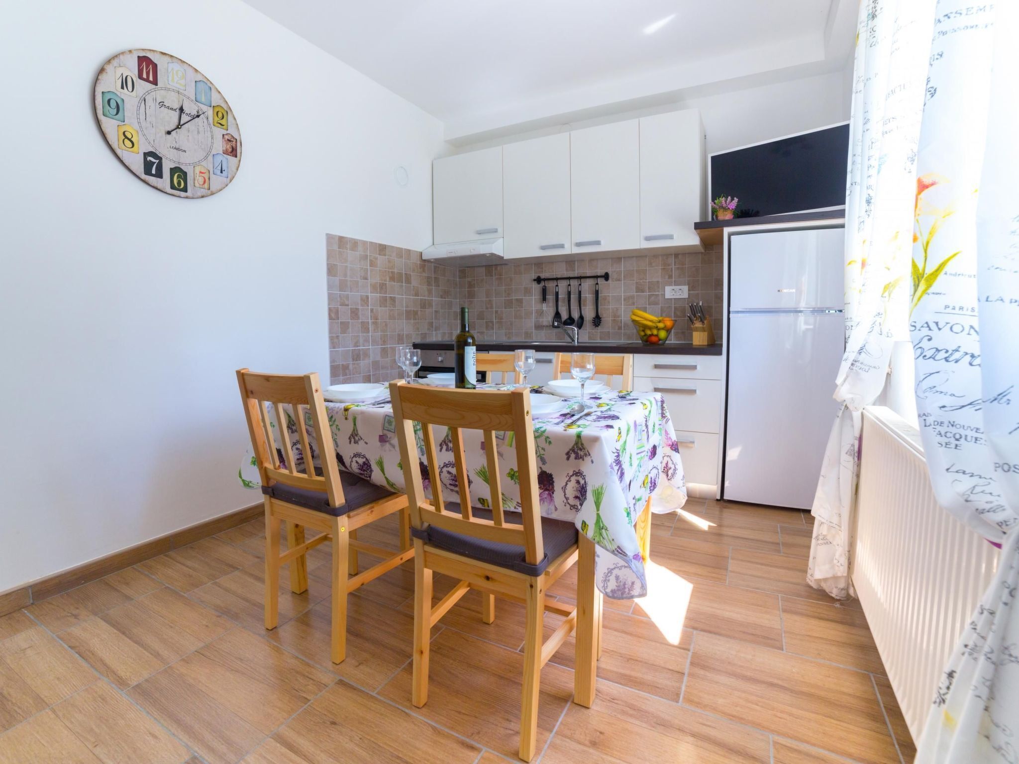 A compact kitchen with white cabinets, a wooden dining table with chairs, and a wall clock.
