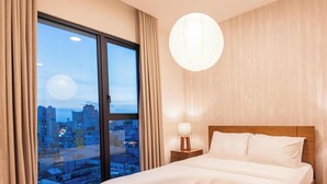 Ett hotellrum med en stor säng, ett nattduksbord, en lampa och utsikt över staden genom ett stort fönster.