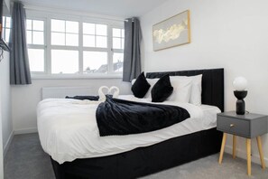 A modern bedroom with a black bed, white bedding, a grey bedside table, and a window with curtains. - Haydock Work Crew Haven Sycamore Avenue Free Parking (St Helens)
