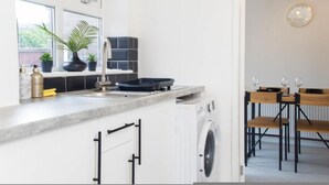 Una cocina moderna con lavadora integrada, un comedor con mesas y sillas de madera, y un amplio ventanal con plantas.
