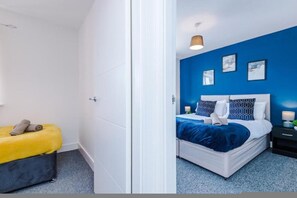 A room with a bed, a bedside table, a nightstand, a yellow bench, and a door. - Penny Bridge Basecamp 6-bed Contractor House St Helens (St Helens)