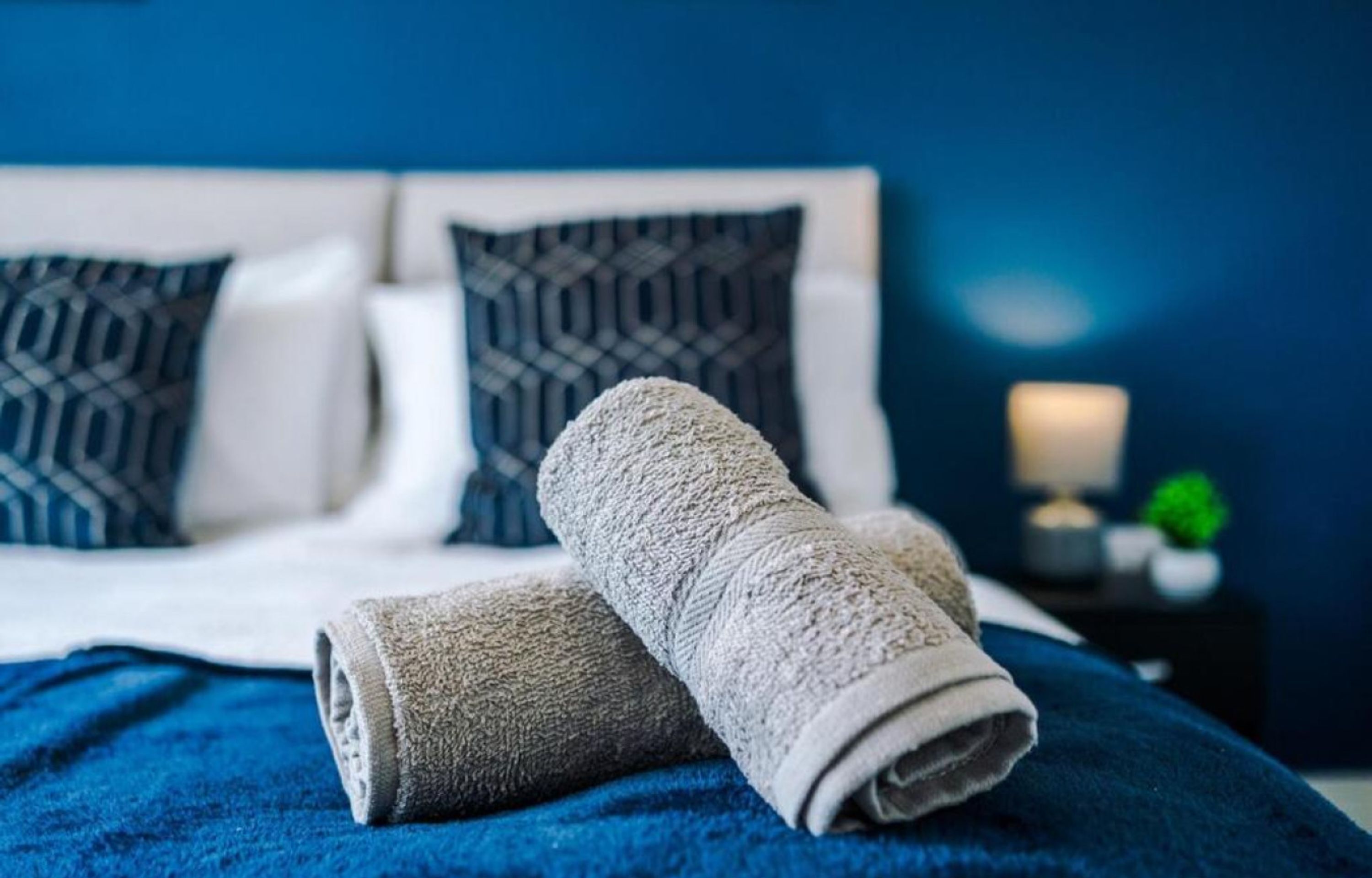 A neatly made bed with a blue blanket and two rolled towels, a white headboard, and patterned pillows.