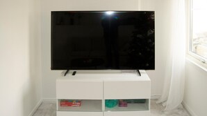 A flat-screen TV on a white stand with a remote control, a glass shelf below holding a few items, and a window with curtains to the right. - Nice home in Brottby with house a panoramic view (Brottby)